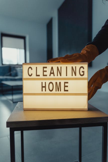 A dustpan and brush set hanging on a light grey wall, with the brush positioned inside the dustpan. The dustpan is made of light blue plastic, and the brush has a wooden handle with soft bristles. The